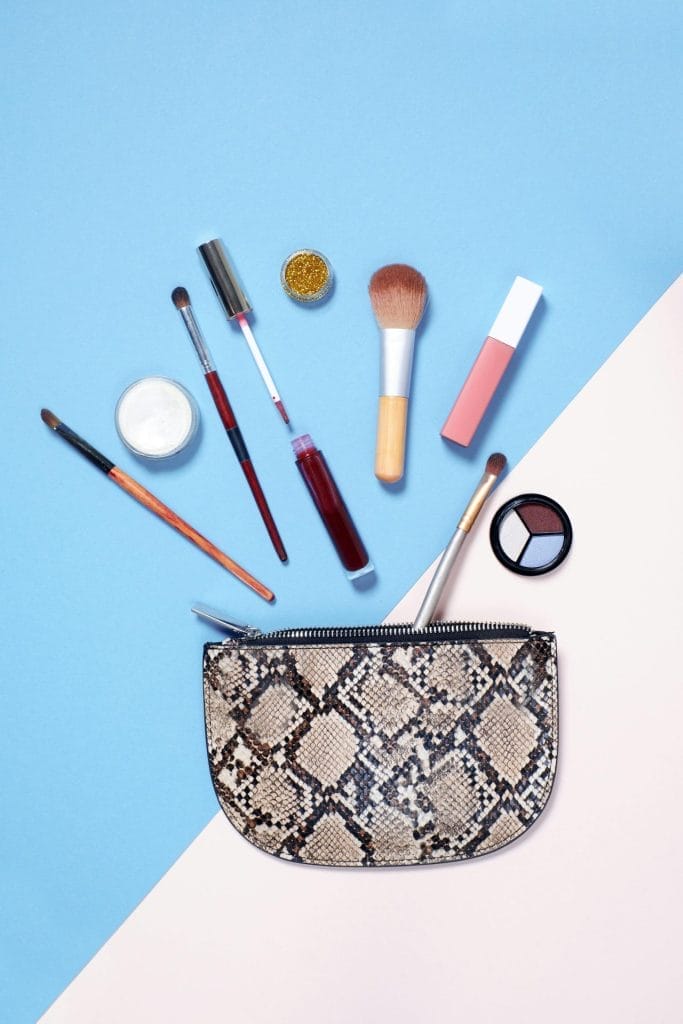 A flat lay of vibrant makeup items arranged around a stylish snakeskin cosmetic bag on a blue and beige background.