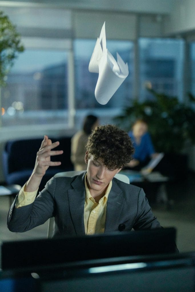 Young office worker in gray suit frustrated, tossing papers indoors.