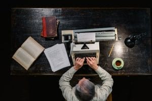 Senior man using typewriter at wooden desk surrounded by books and tea.